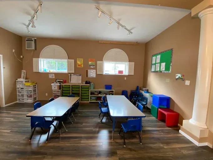 A room with two arched windows, two long tables with small chairs and cubby shelves along the wall.