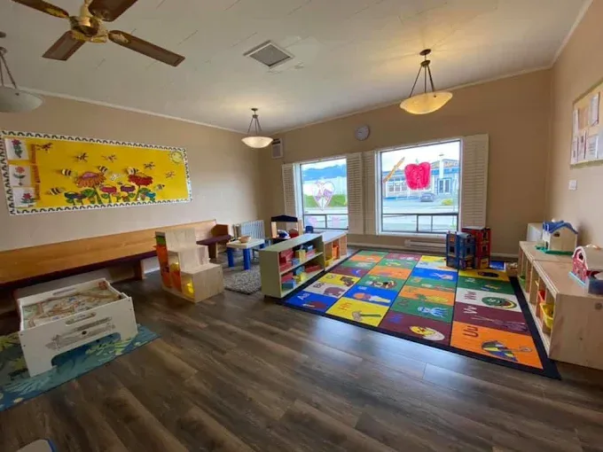 A bright playroom with big front windows, a long wooden bench, low shelves and a colourful alphabet carpet.