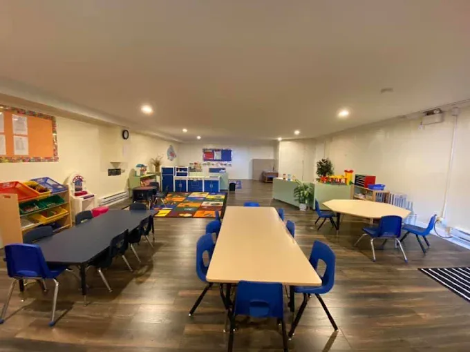 A long room with child-sized tables and blue chairs, low toy shelves and a play kitchen at the far end.
