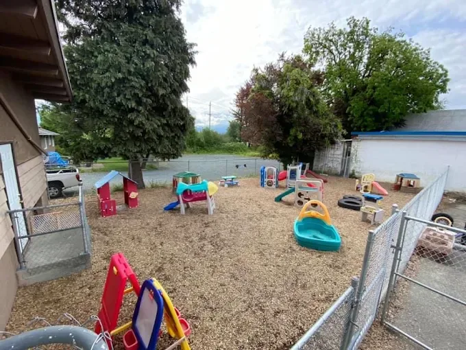 The fenced outdoor yard with wood-chip ground, slides, playhouses, a sandbox and tall trees.
